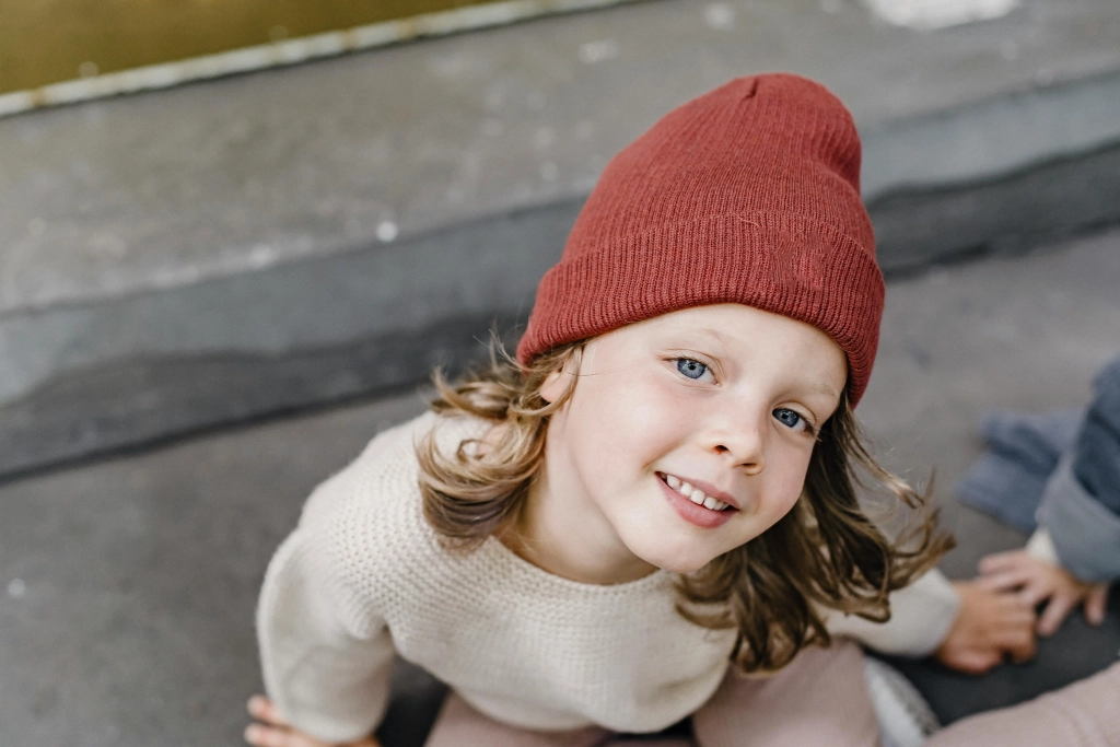 Young child with blond hair and blue eyes, wearing a red hat and white sweater, sitting on pavement in Cochrane and Calgary, looking thoughtfully at the camera.